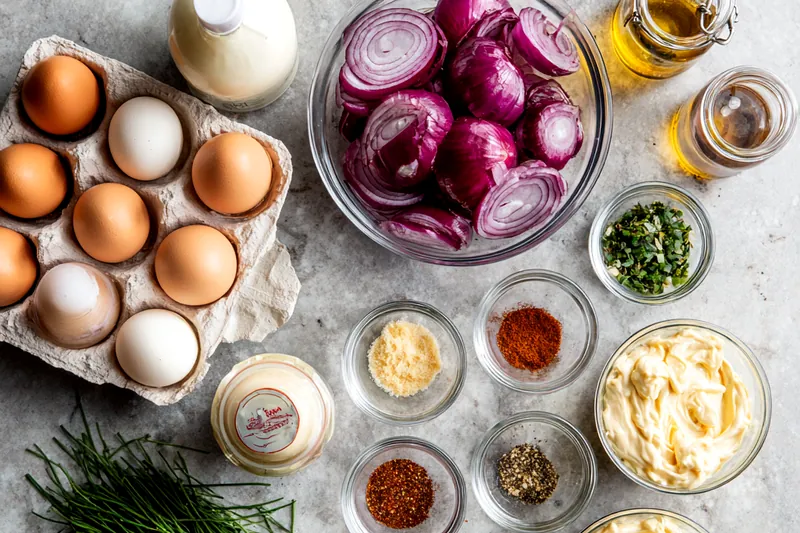 Raw ingredients for deviled eggs including carton of eggs, red onion slices, mayonnaise, mustard, vinegar, and spices on a marble counter